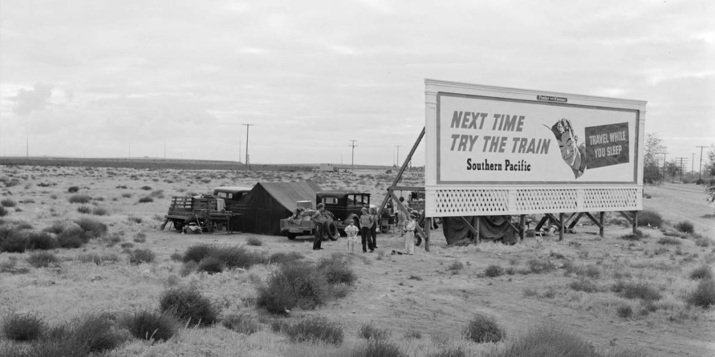 "Three families camped on the plains along U.S. 99 in California. They are camped behind a billboard which serves as a partial windbreak. All are in need of work." 1938. Billboard reads Next Time Try The Train. Southern Pacific. Travel While You Sleep