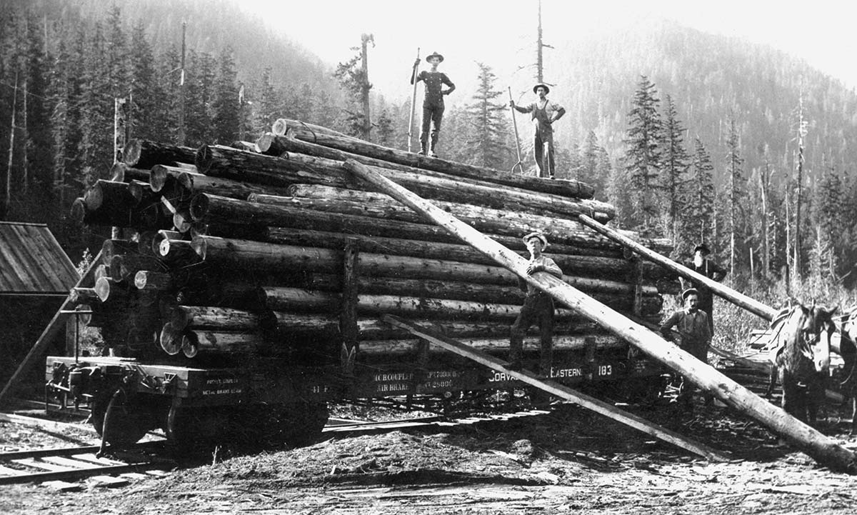 Historic photograph of loggers loading large timber onto a railcar, representing early 20th-century logging and rail transport common during the Depression era.